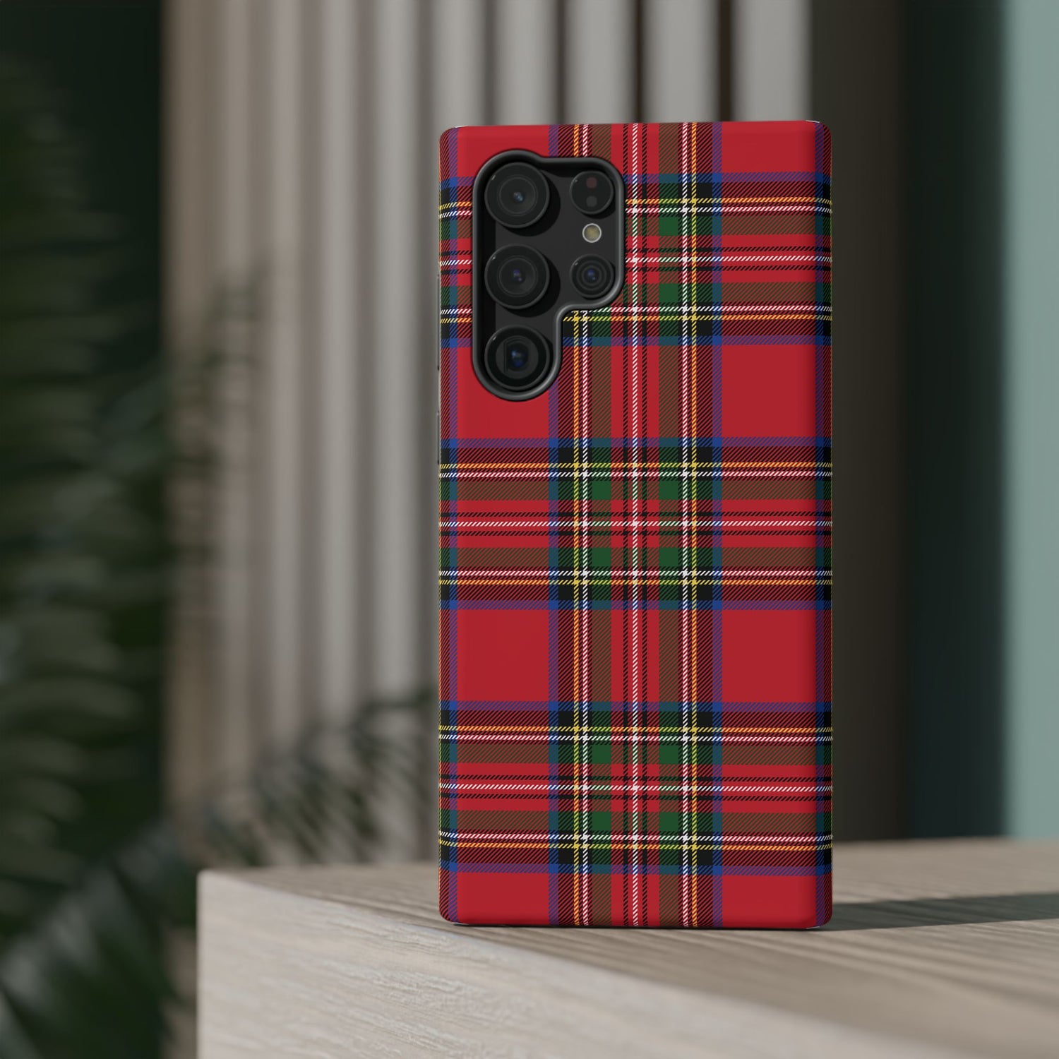 A red and green tartan plaid patterned plastic phone case with a camera module on the back, placed on a wooden surface with a blurred background.